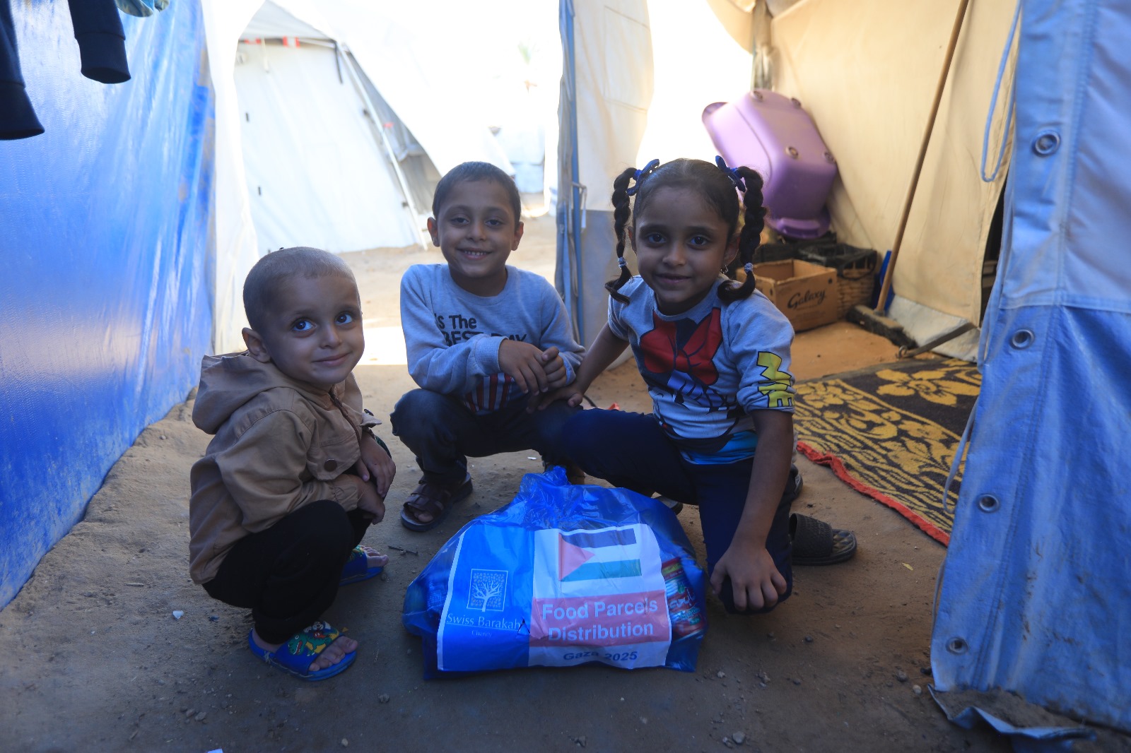 Three children smile while sitting next to a blue food parcel inside a humanitarian tent.