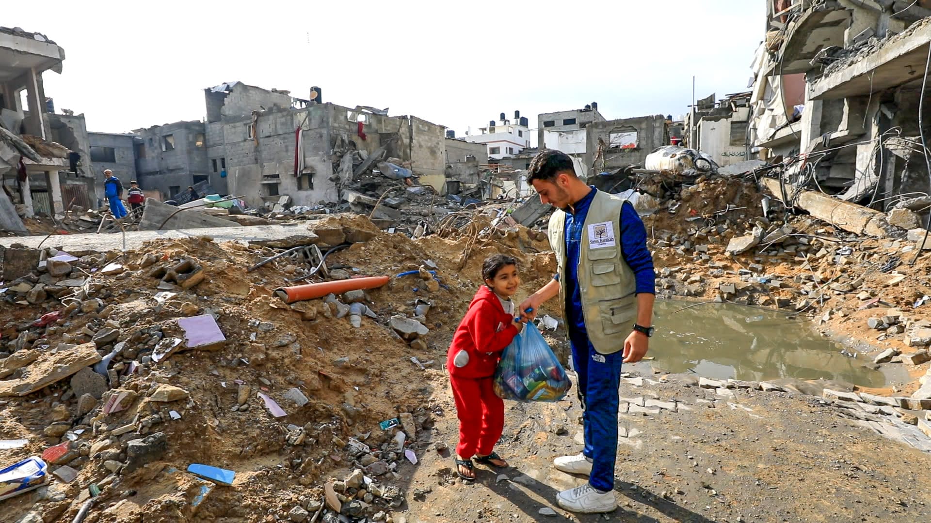 Un travailleur humanitaire remet un sac de nourriture à une jeune fille souriante en survêtement rouge au milieu de bâtiments détruits.