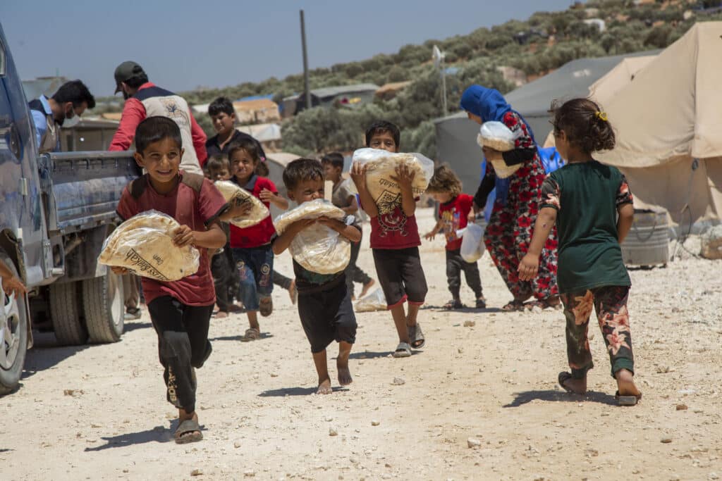 Un groupe d'enfants sourit et court dans un camp de réfugiés en portant de grands sacs de pain plat frais provenant d'une distribution de Swiss Barakah Charity.