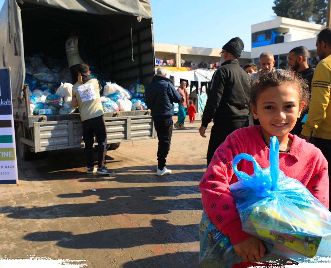 A young girl smiles while carrying a blue plastic bag filled with food aid near a humanitarian supply truck.