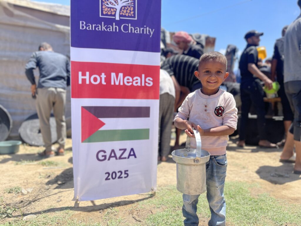 A smiling boy holds a metal bucket for food next to a "Hot Meals Gaza 2025" banner from Barakah Charity.